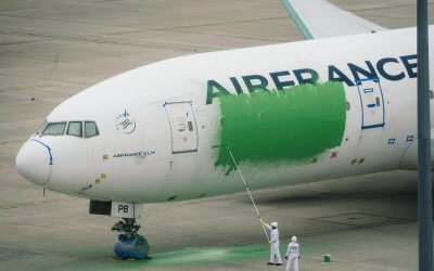 Dos personas con trajes blancos pintan de verde un avión comercial de la aerolínea Air France, cubriendo parte del fuselaje en una pista de aeropuerto.