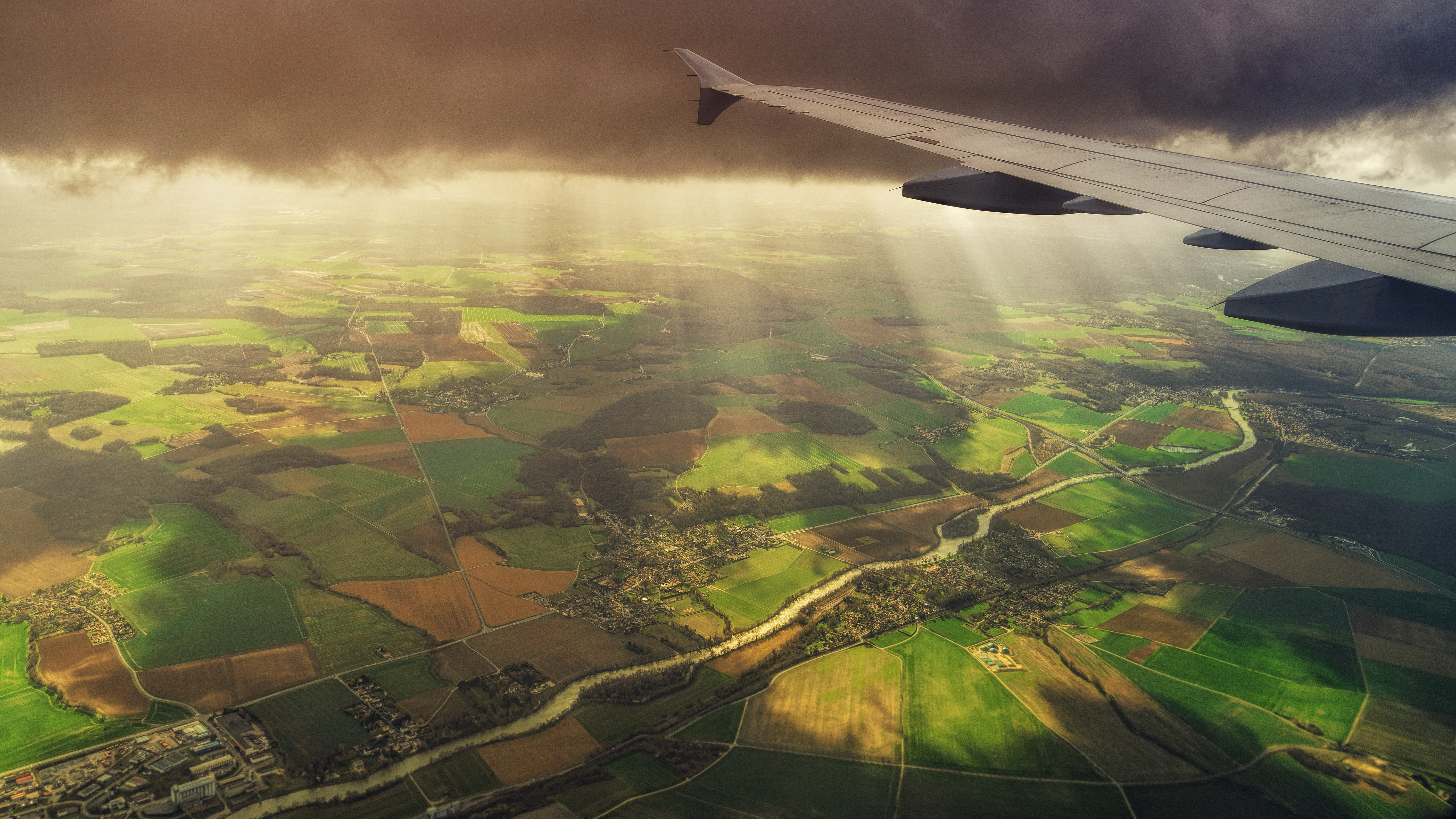 Vista aérea de campos verdes y marrones iluminados por la luz solar a través de nubes oscuras, captada desde la ventana de un avión en vuelo.