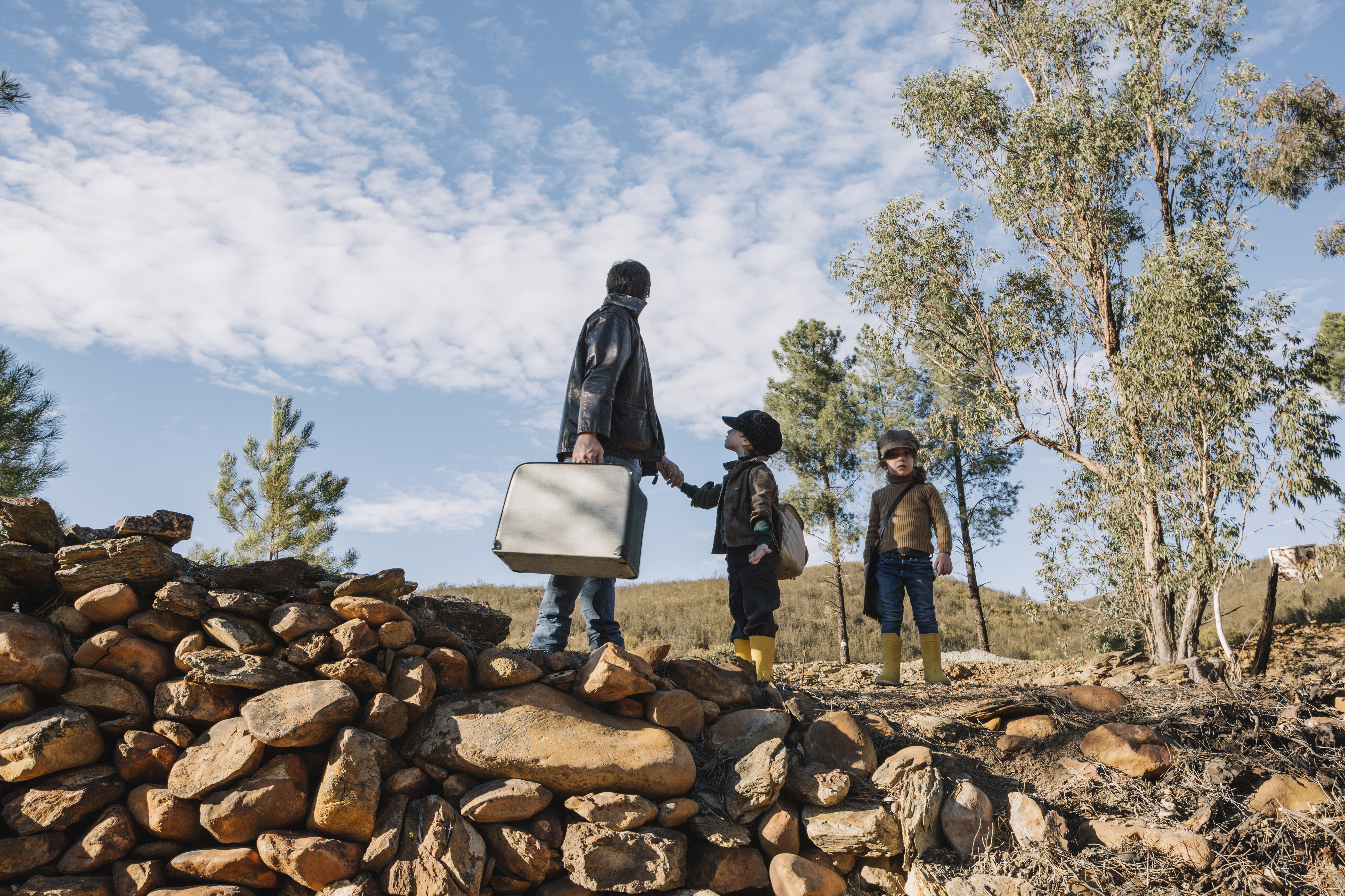 Padre con dos niños caminando por una zona rural de Pinamar con una valija, en un paisaje natural con árboles y cielo despejado.