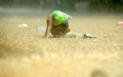 Persona caminando entre aguas marrones por una inundación, cubierto hasta los hombros, mientras sostiene provisiones sobre su cabeza bajo la lluvia intensa.