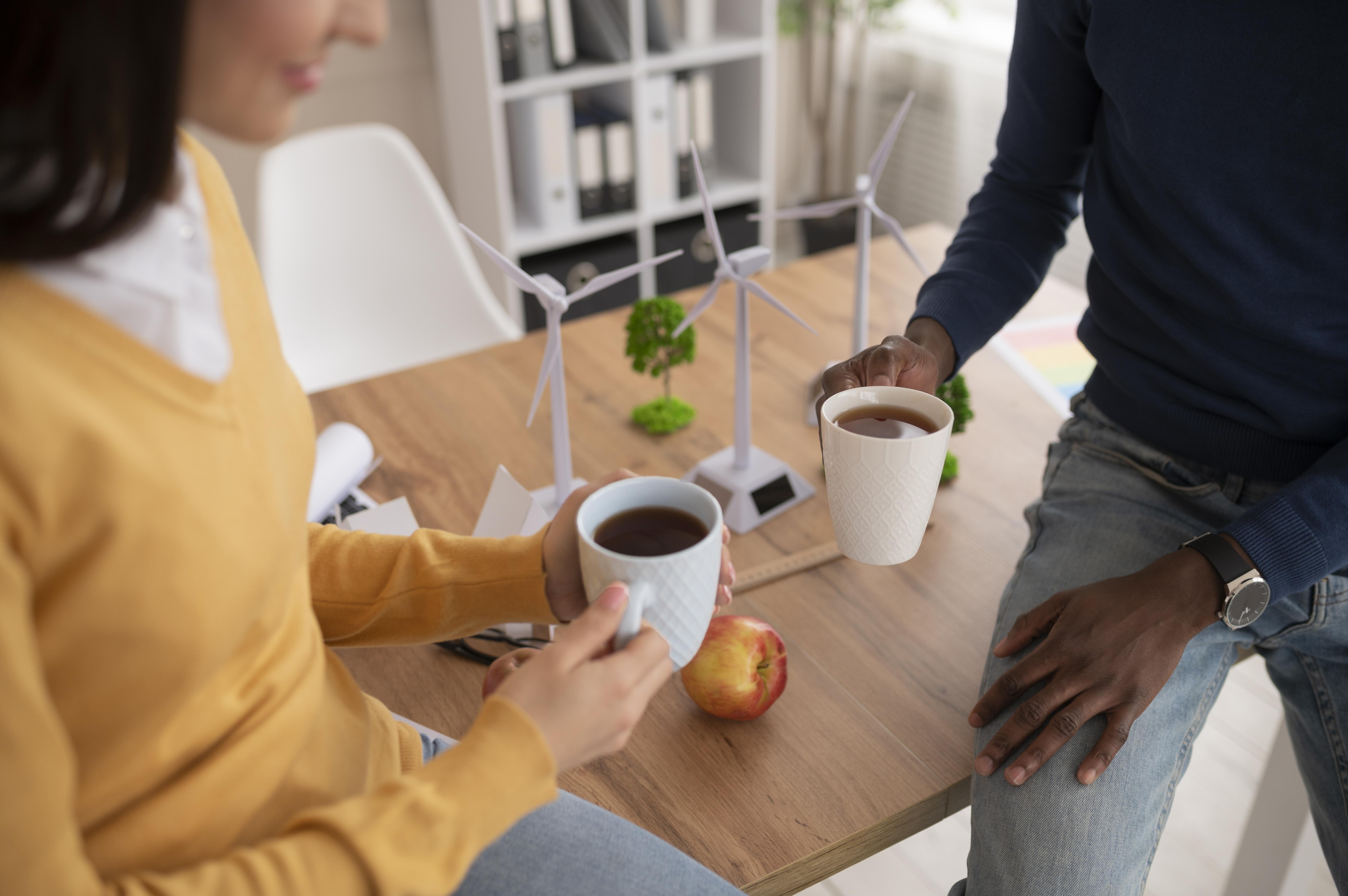 Dos personas conversan mientras toman café frente a una maqueta de turbinas eólicas, en un entorno laboral vinculado a energías renovables.