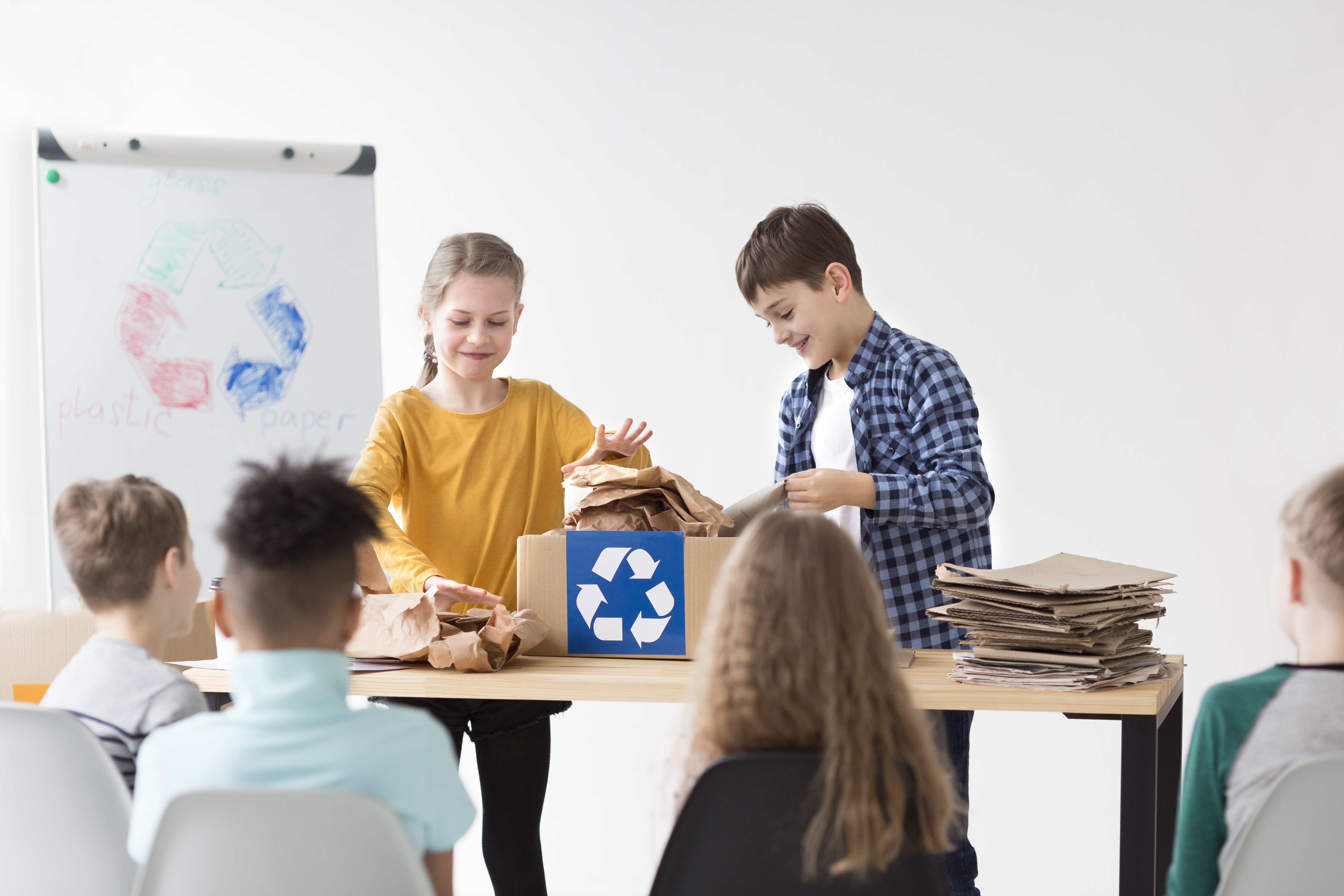 Niños y niñas en un aula escolar reciclan papel mientras aprenden sobre sostenibilidad, rodeados de carteles de separación de residuos.
