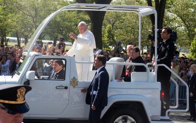 Papa Francisco de pie en el papamóvil saludando a la multitud en una calle arbolada.