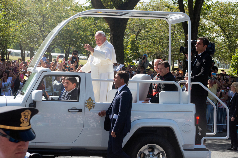 Papa Francisco de pie en el papamóvil saludando a la multitud en una calle arbolada.