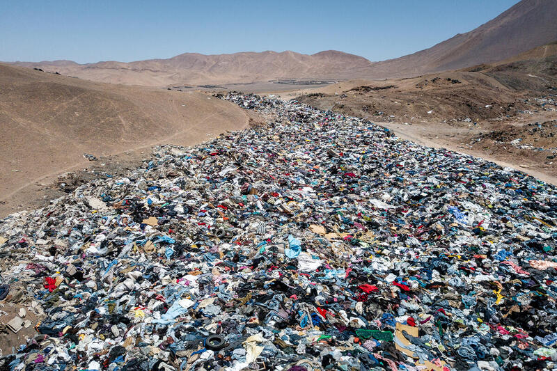 Gran acumulación de ropa usada en un vertedero textil ubicado en el desierto de Atacama, rodeado de montañas áridas y cielo despejado.