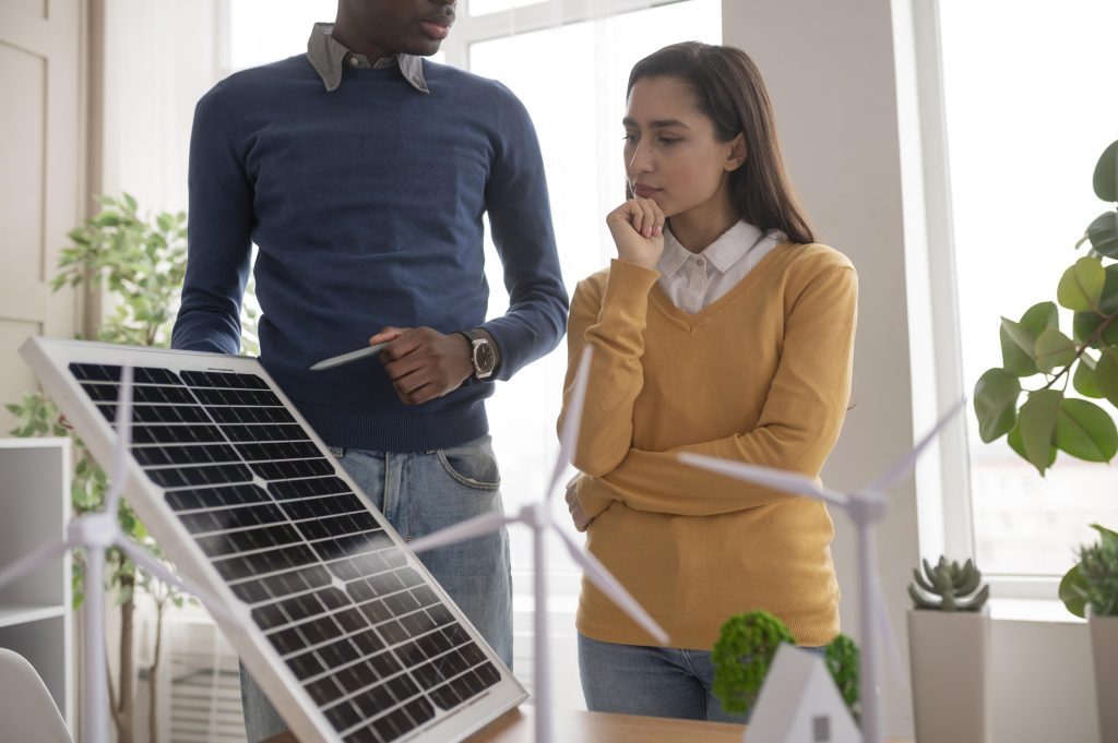 Dos personas analizan un panel solar junto a modelos de turbinas eólicas en una oficina sostenible.