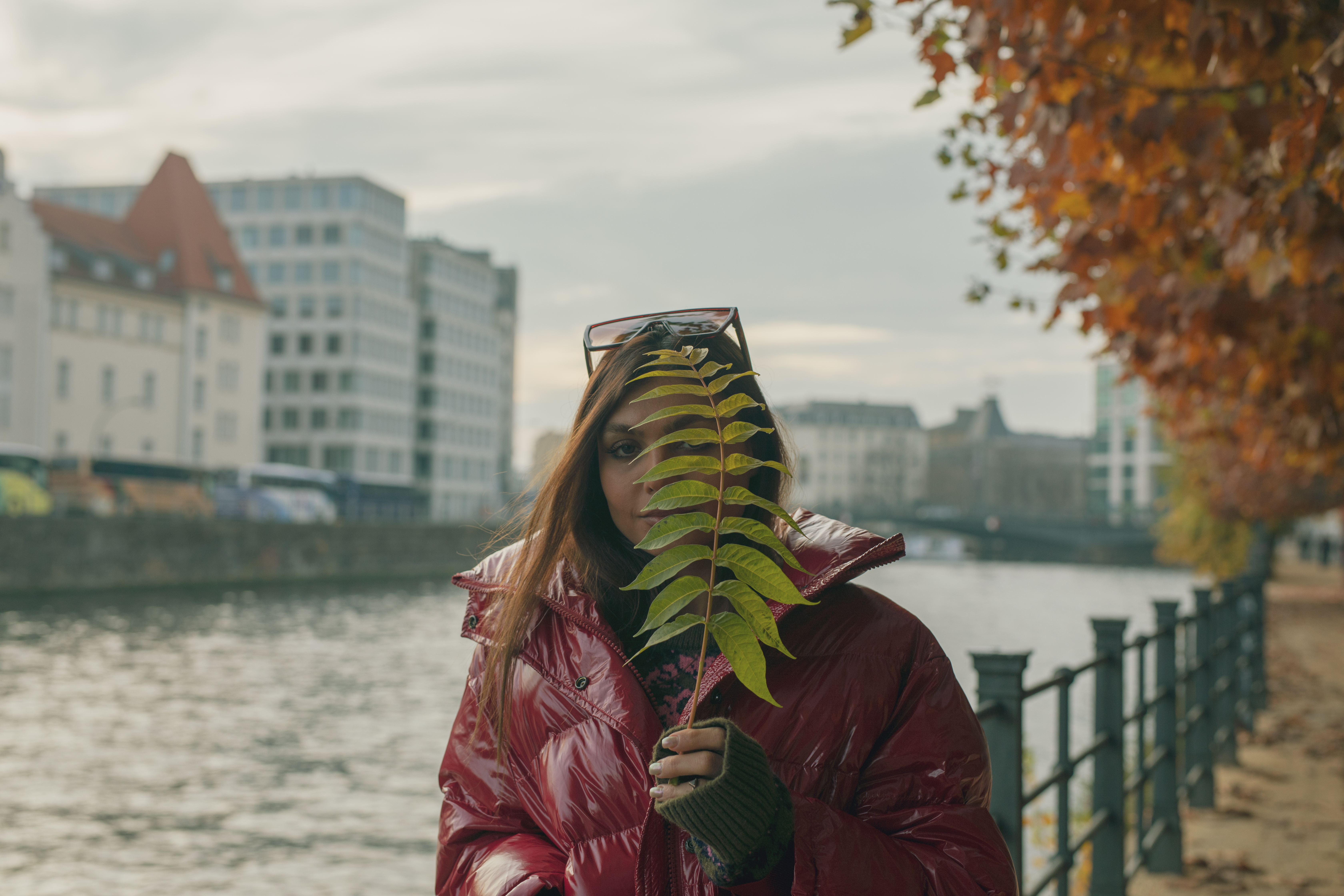 Mujer con campera burdeos tapa parte de su rostro con una hoja frente a un canal en otoño.
