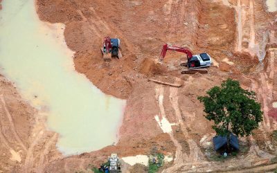 Vista aérea de un terreno de excavación con dos retroexcavadoras operando cerca de un estanque de agua turbia, un árbol aislado y una carpa negra en la parte inferior.