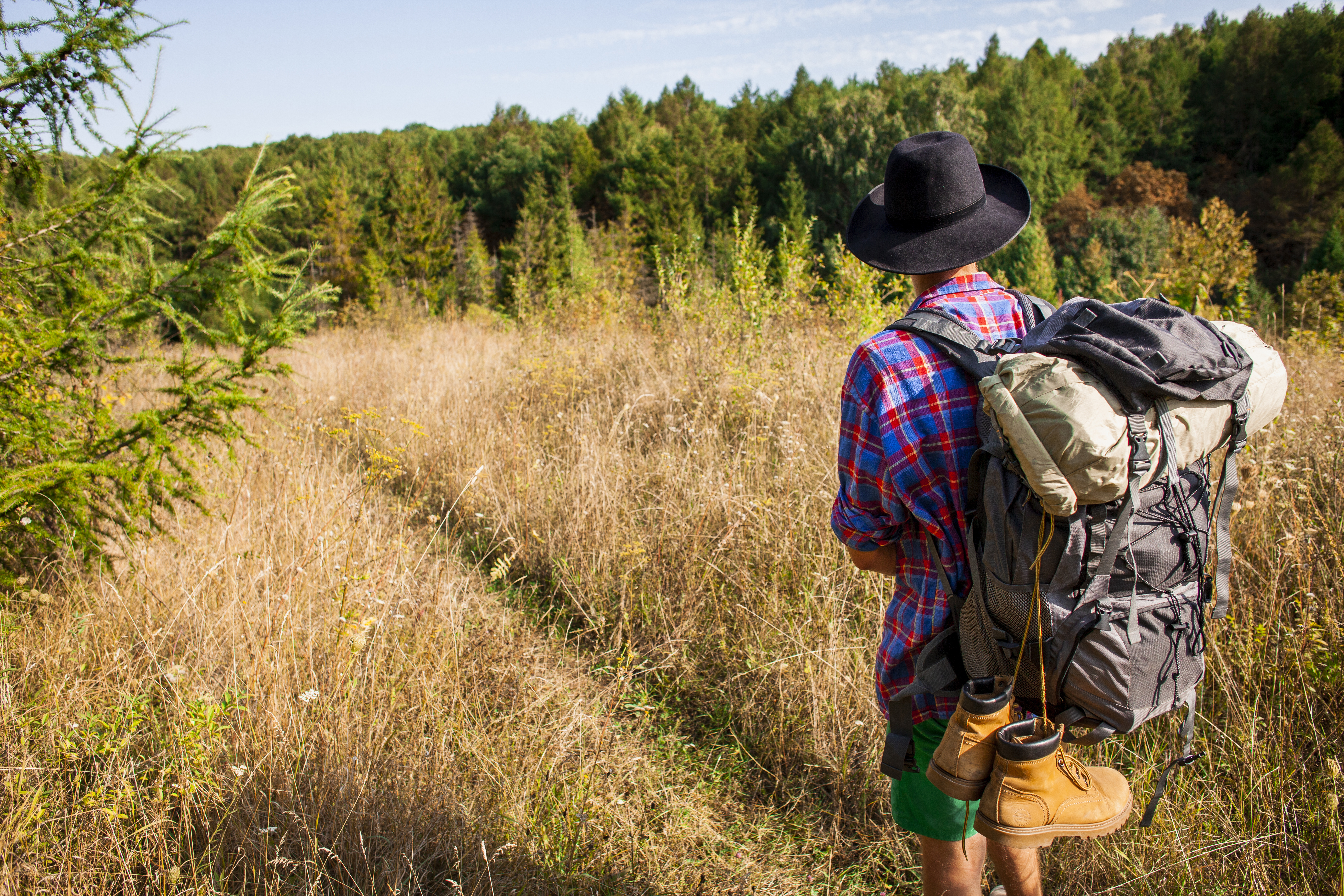 Un hombre con camisa a cuadros, sombrero negro y mochila de campamento avanza por un sendero rodeado de pastizales secos y árboles frondosos. Lleva un par de botas atadas a su mochila, lo que sugiere una larga travesía. La imagen transmite conexión con la naturaleza, aventura y exploración al aire libre.