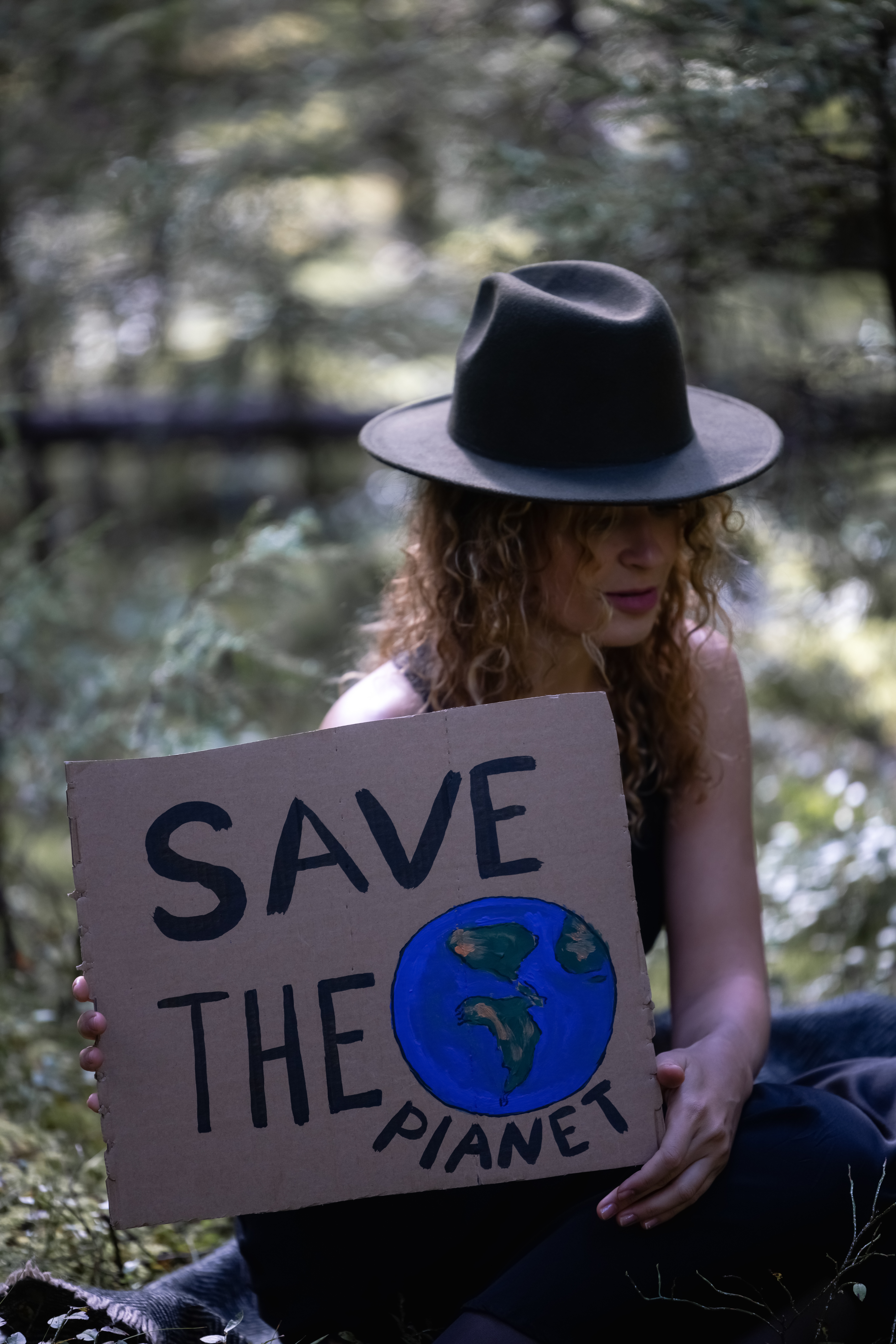 Mujer con sombrero en el bosque sostiene cartel que dice “Save the planet” con dibujo del planeta Tierra.