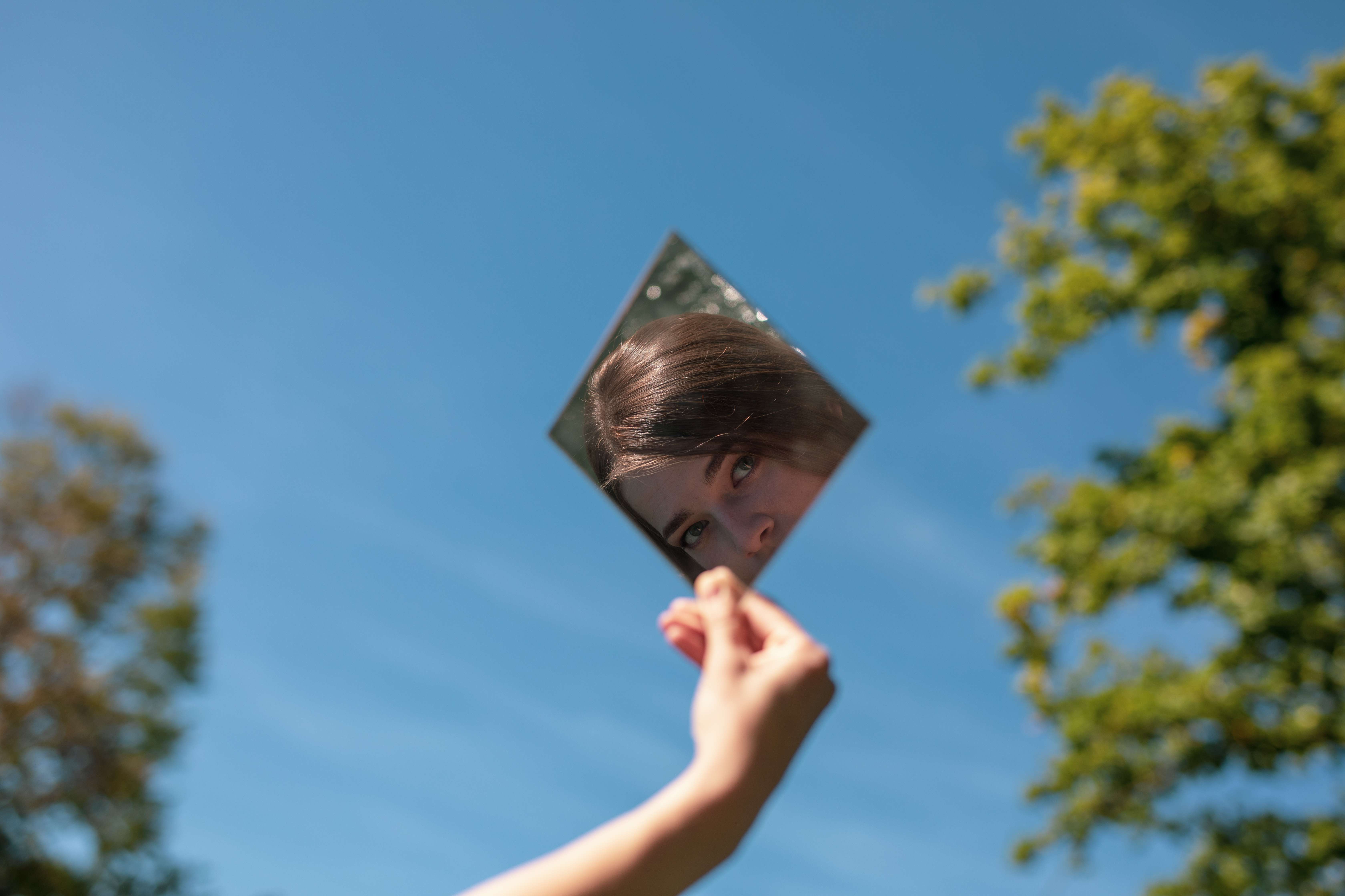 Mano sosteniendo un espejo con el reflejo de una mujer joven contra un cielo azul.
