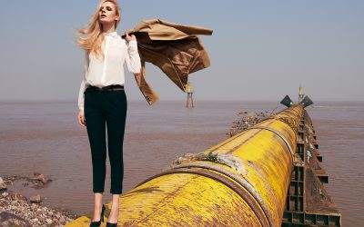 Mujer rubia con camisa blanca y pantalón negro de pie sobre una gran tubería amarilla junto al mar, sosteniendo un abrigo beige al viento.