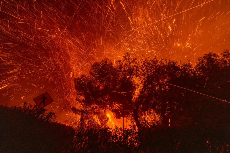 Incendio forestal nocturno con llamas intensas y chispas elevándose entre los árboles, iluminando el cielo en tonos rojos y anaranjados.