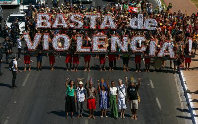 Manifestación indígena en una avenida con personas sosteniendo un gran cartel que dice “BASTA DE VIOLÊNCIA”, acompañadas por líderes comunitarios con los brazos en alto.