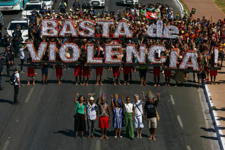 Manifestación indígena en una avenida con personas sosteniendo un gran cartel que dice “BASTA DE VIOLÊNCIA”, acompañadas por líderes comunitarios con los brazos en alto.
