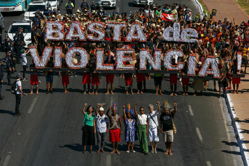 Manifestación indígena en una avenida con personas sosteniendo un gran cartel que dice “BASTA DE VIOLÊNCIA”, acompañadas por líderes comunitarios con los brazos en alto.