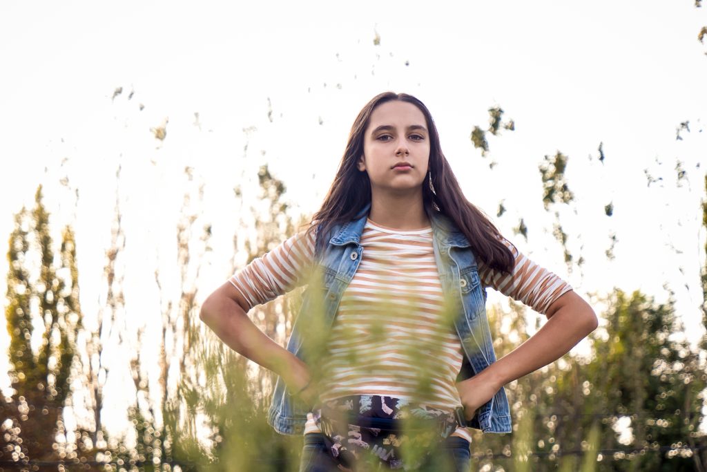 Retrato de una joven de pie al aire libre, con las manos en la cintura y expresión decidida, rodeada de vegetación y luz natural.