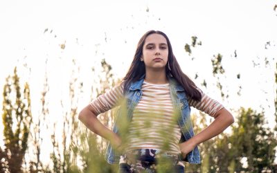 Retrato de una joven de pie al aire libre, con las manos en la cintura y expresión decidida, rodeada de vegetación y luz natural.